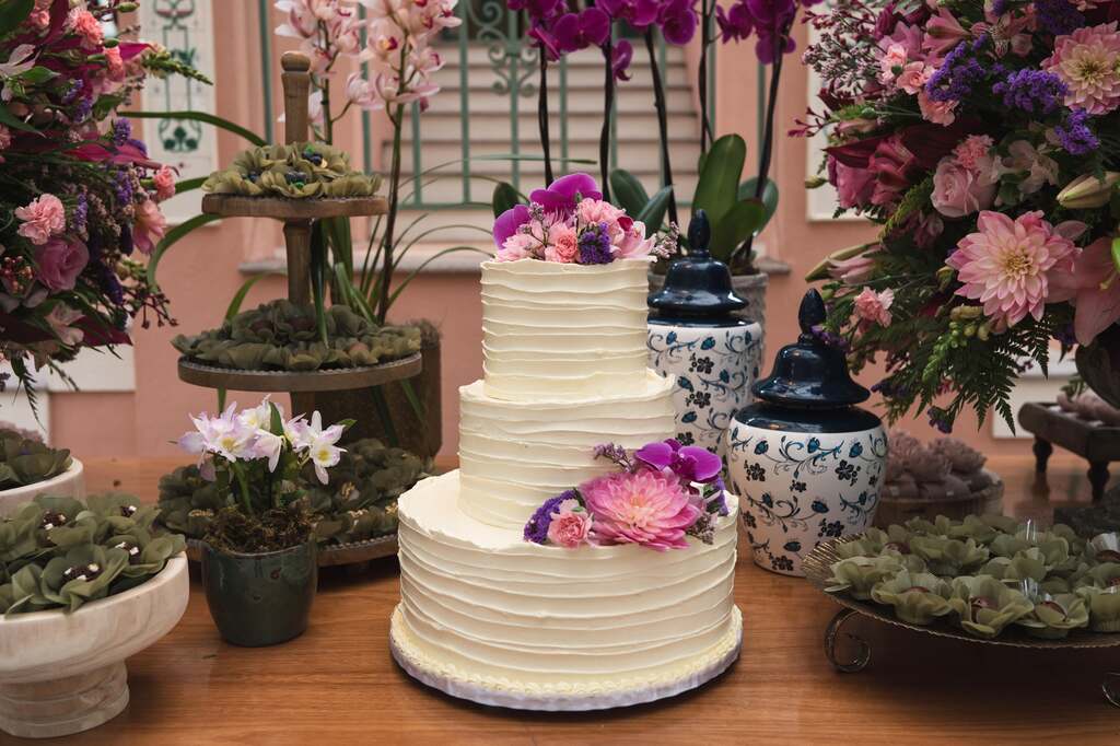 bolo de casamento branco com três andares com flores rosas e roxas no topo e ao lado com bandejas de madeira com doces de casamento com forminhas de flores verdes