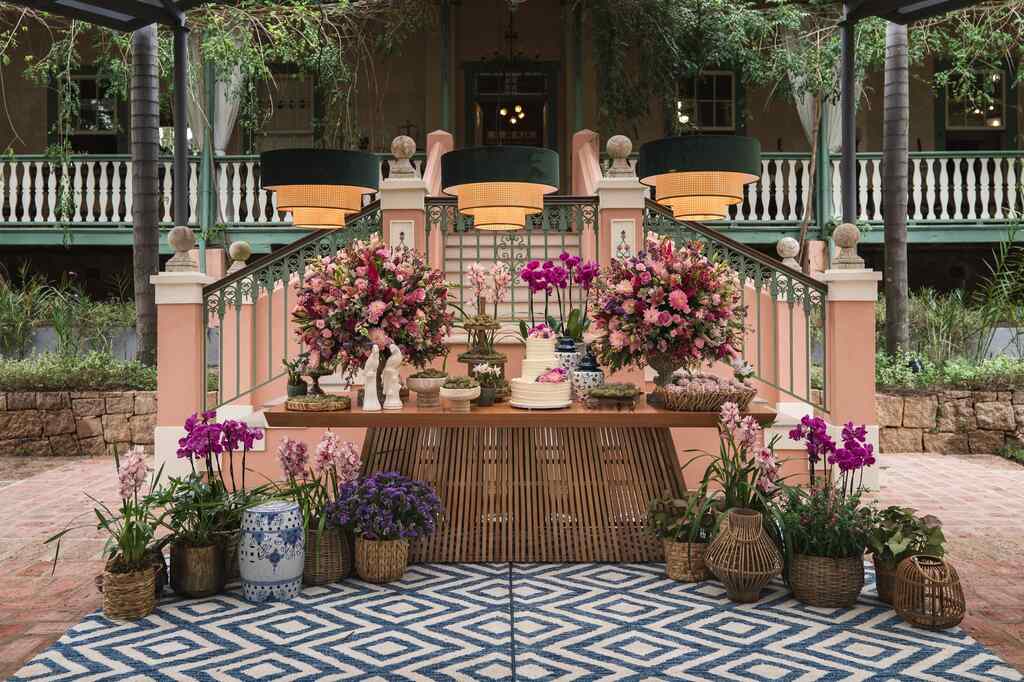 mesa de madeira com dois vasos com flores rosas e roxas e bolo de casamento branco com três andares e vasos de palha no chão com flores rosas roxas e lilás