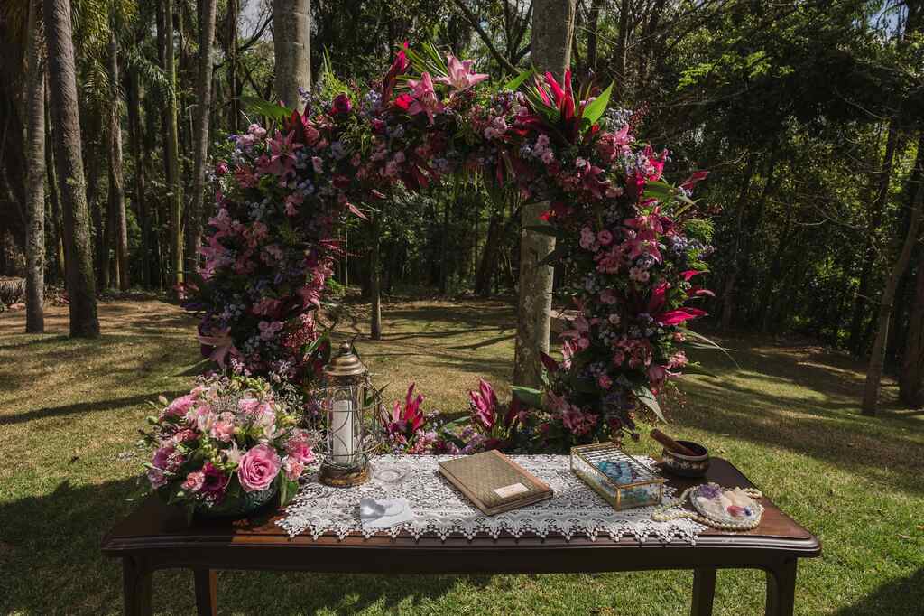 altar redondo com flores rosas roxas e liláses no jardim e mesa de madeira com toalha bordada na frente