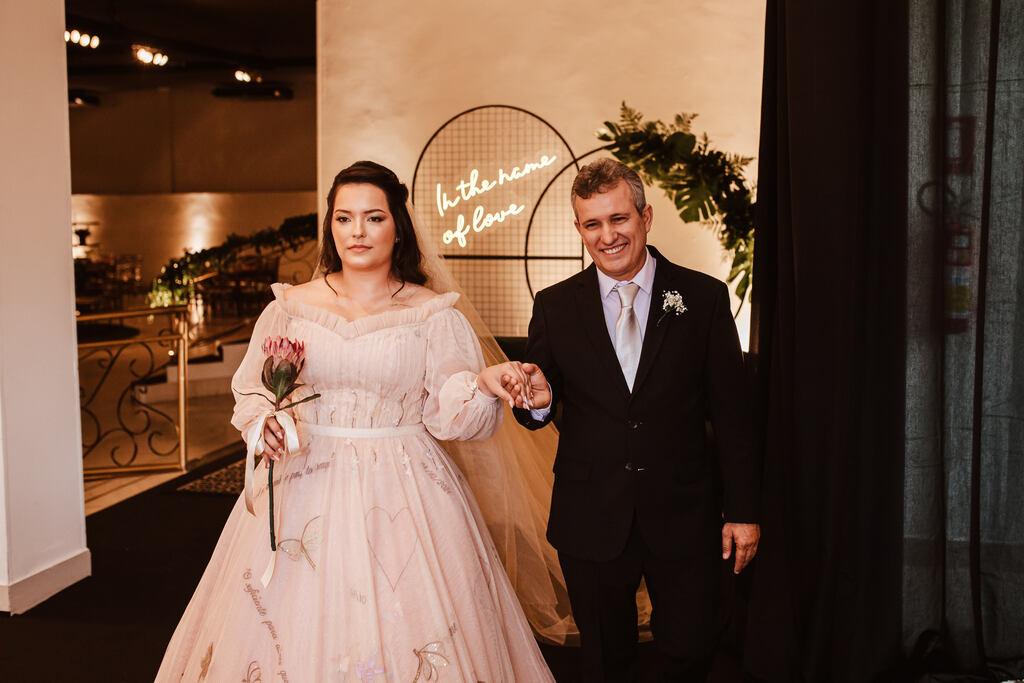 noiva com vetsido rosÊ com manga longa segurando flor protea de mãos dadas com o pai com tenro preto