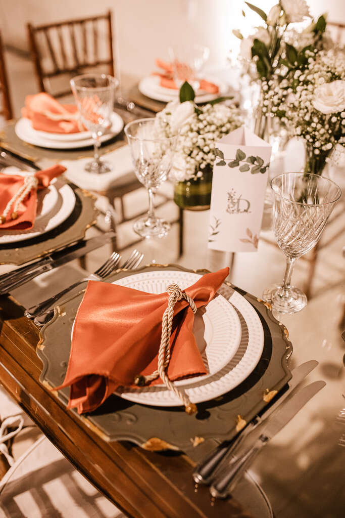 mesa de vidro com lugar americano verde e mesa posta com guardanapo laranja e flores brancas no centro da mesa