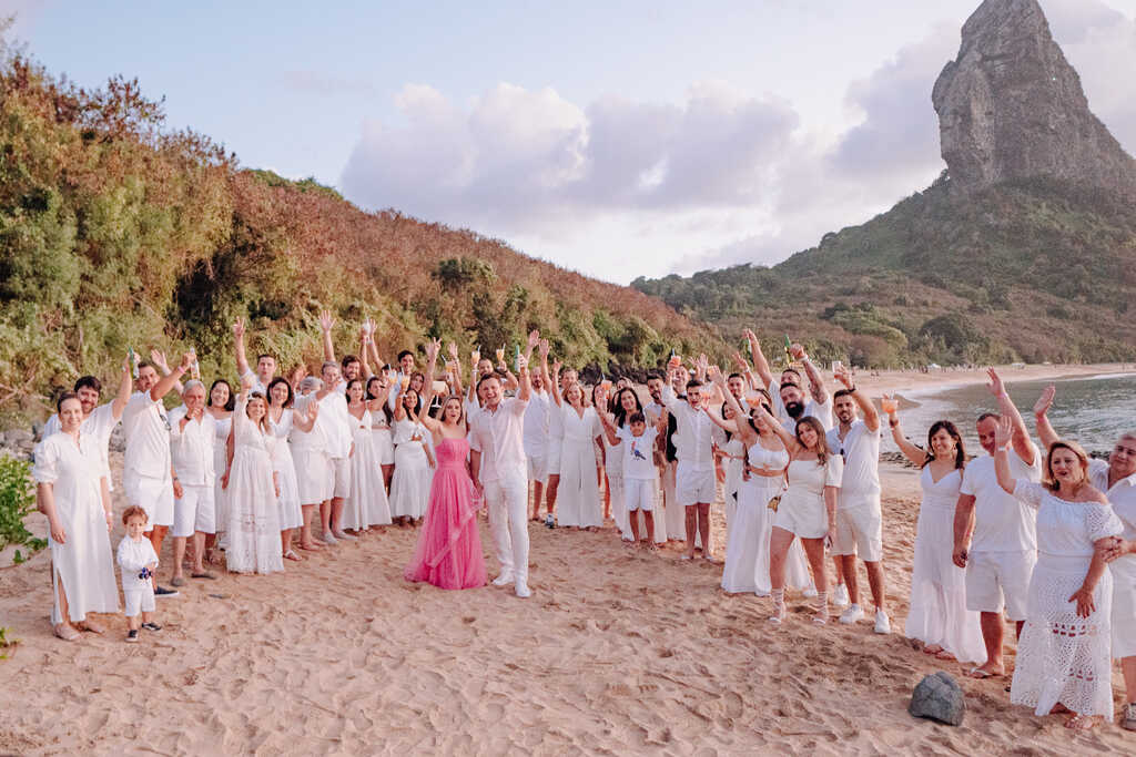 noiva com vetsido rosa longo ao lado do noivo com roupa branca na praia e convidados com roupas brancas na praia de fernando de noronha