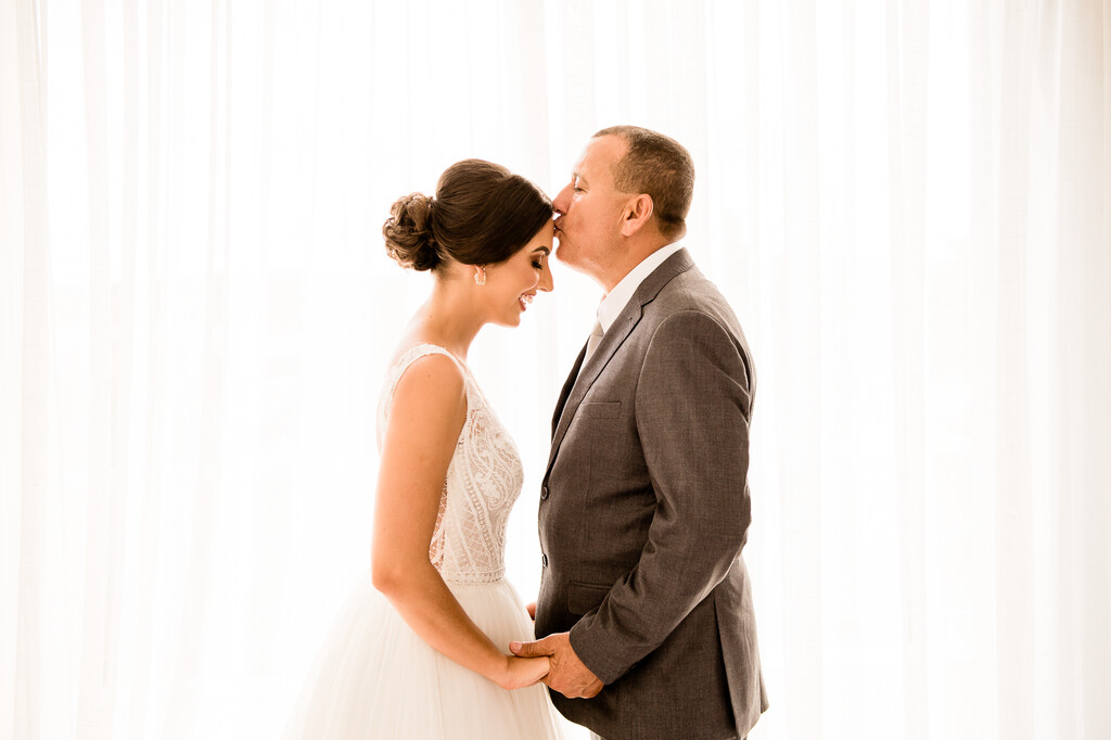pai da noiva com terno cinza de mãos dadas com a noiva beijando a testa dela