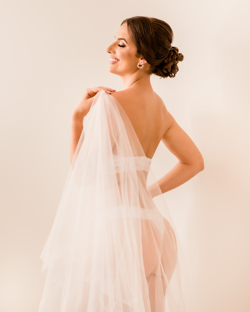 mulher de lingerie de costas segurando véu de noiva em ensaio boudoir