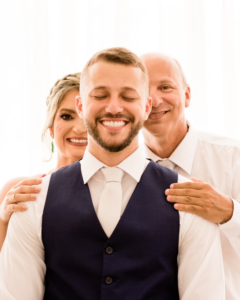 pais do noivo segurando nos ombros do noivo com camisa e gravata branca com colete azul marinho