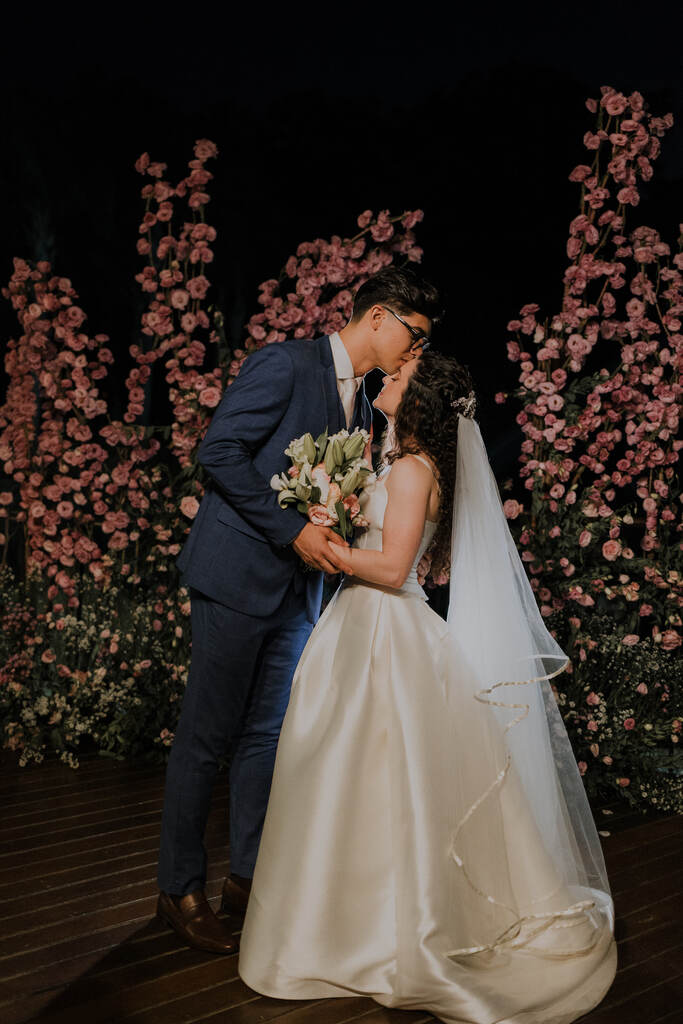 noivo com terno azul beijando a testa da noiva em deck de madeira decorado com flores cor de rosa