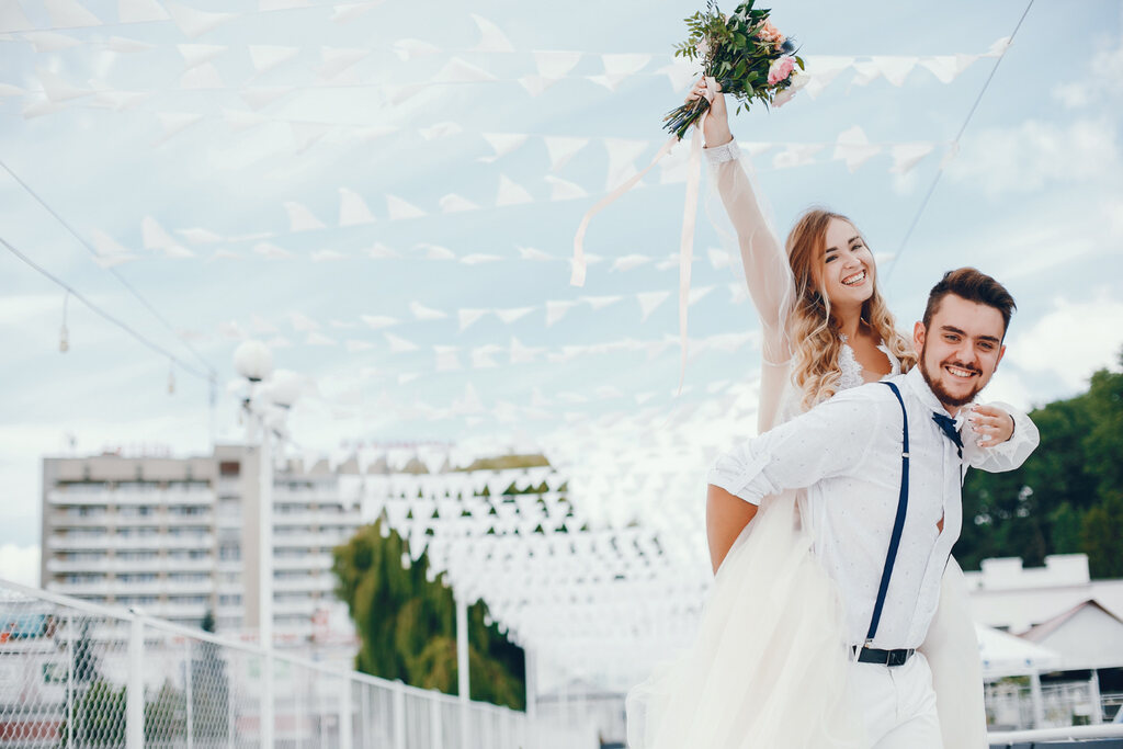 noivo feliz com camisa branca supensório e gravata borboleta azul carregando noiva no colo erguendo buquê ao ar livre