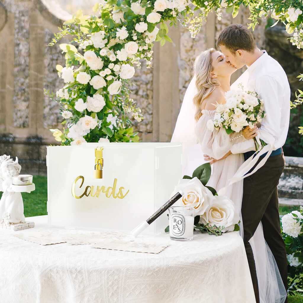 noivos se beijando ao fundo e caixa semi transparente com cadeado e escrito cards em dourado em cima da mesa para arrecadar dinheiro para casamento
