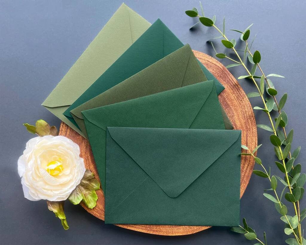 envelopes para carta em diferentes tons de verde sobre mini tapete marrom e ao lado de flor branco e ramos de planta