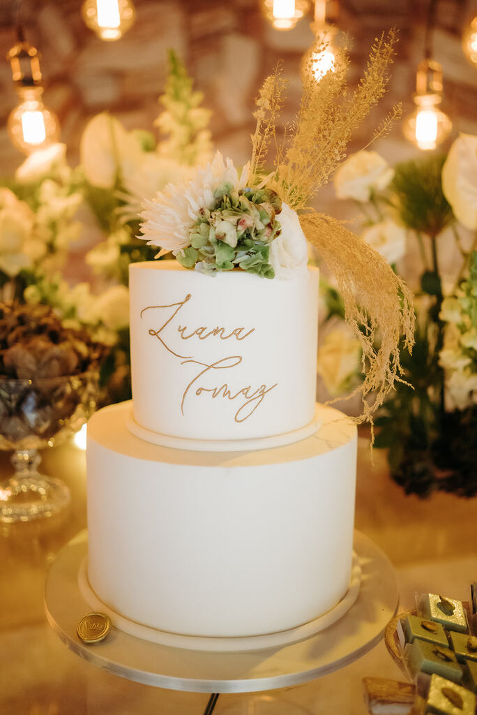 bolo de casamento branco com dois andares com nome dos noivos escrito no bolo e topo com flores brancas e planta