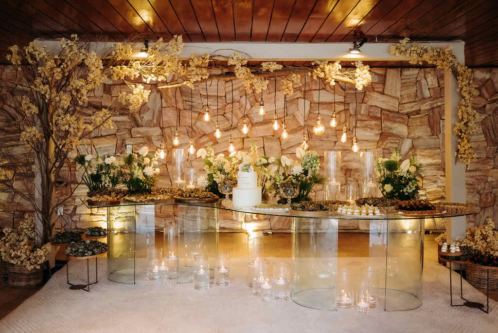 salão com mesa de vidro com bolo de casamento branco ao lado de arranjos com flores e luminarias pendentes