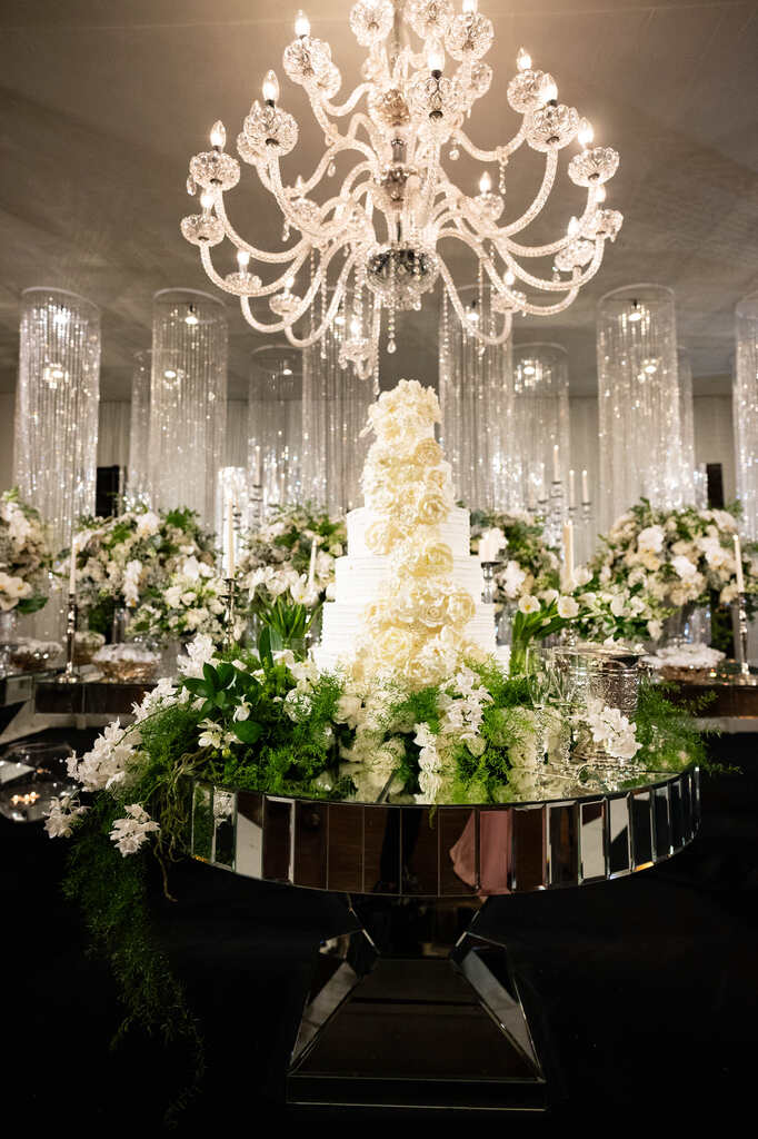 salão com mesas espelhadas com bolo de casamento branco com topo de flores