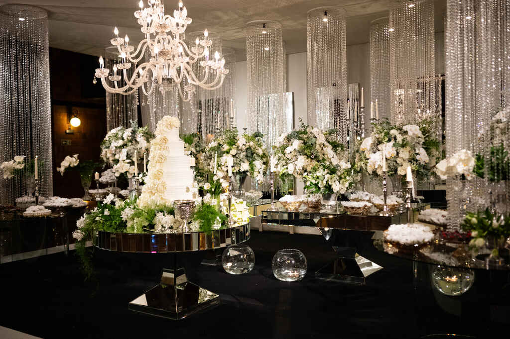 salão com mesas espelhadas com bolo de casamento branco com topo de flores 