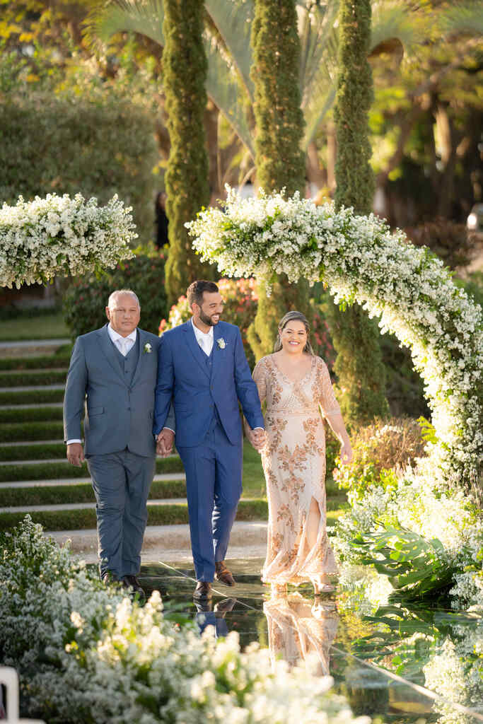 entrada do novo com terno azul com colete de mãos dadas com o pai de terno cinza com colete e a mãe com vestido nude e rosa