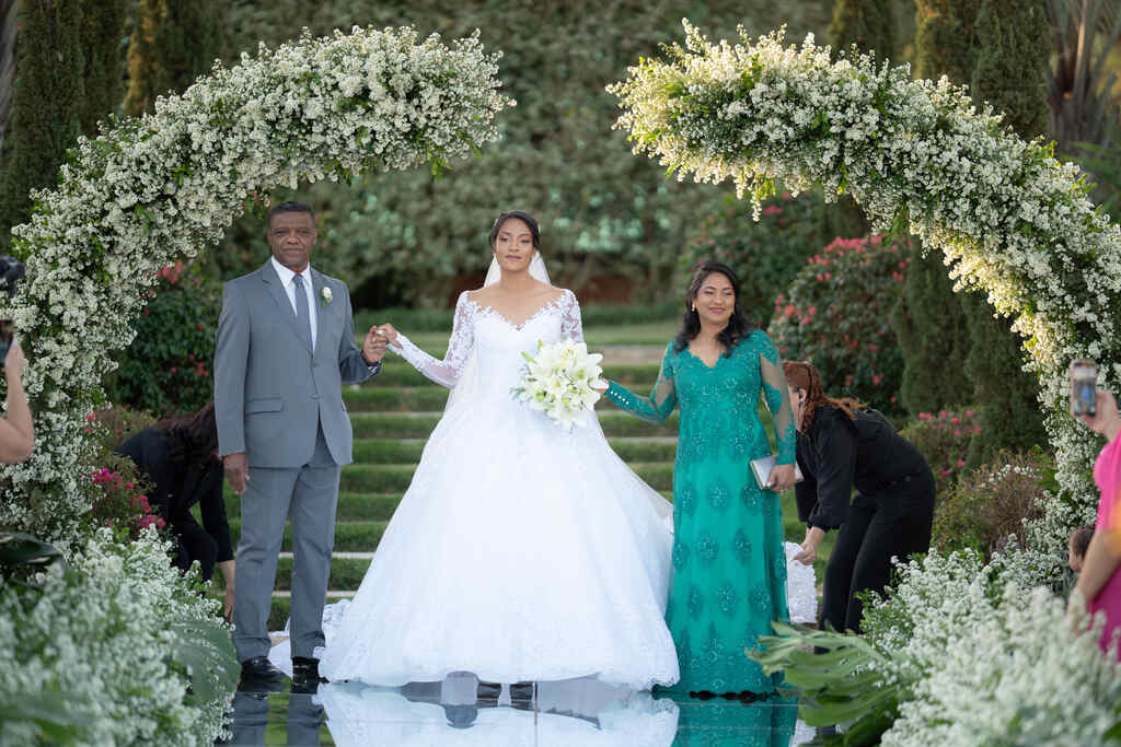 entrada da noiva com vestido com saia volumosa de mãos dadas com o pai com terno cinza com gravata e mãe com vestido verde