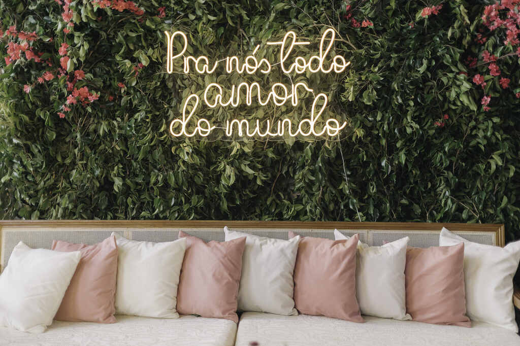 sofá branco com almofadas brancas e rosas e atrás um painel com plantas e flores rosas com letreiro neon