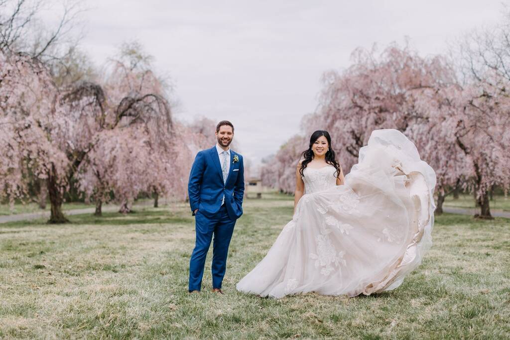 noivo com terno azul ao lado da noiva com vestido com cauda volumosa em meio a jardim com cerejeiras ao fundo