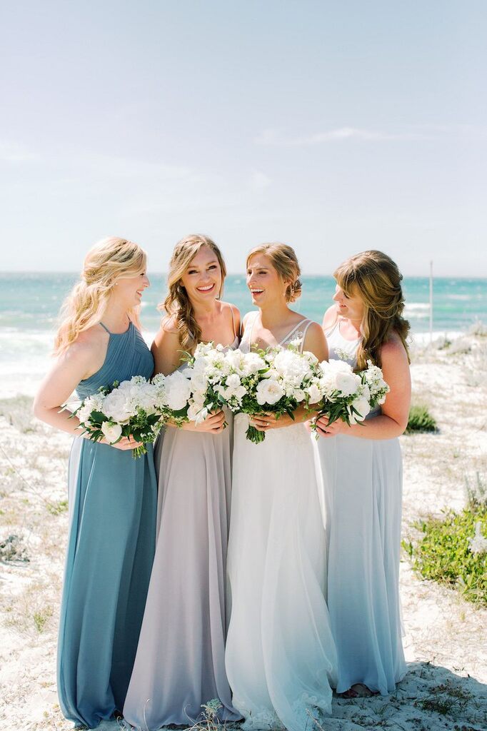 noiva ao lado das madrinhas com vetsidos azuis longos e todas segurando buquês com flores brancas na praia