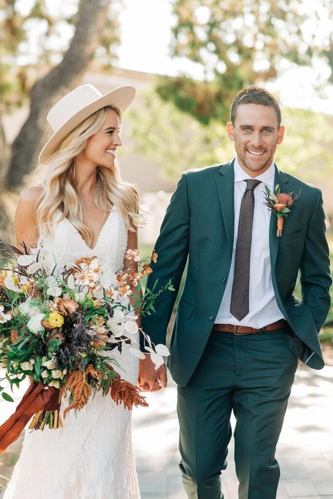 noivo com terno verde com gravata slim e lapela com flores laranjas de mãos dadas com noiva com chapéu de palha e buquê com flores coloridas
