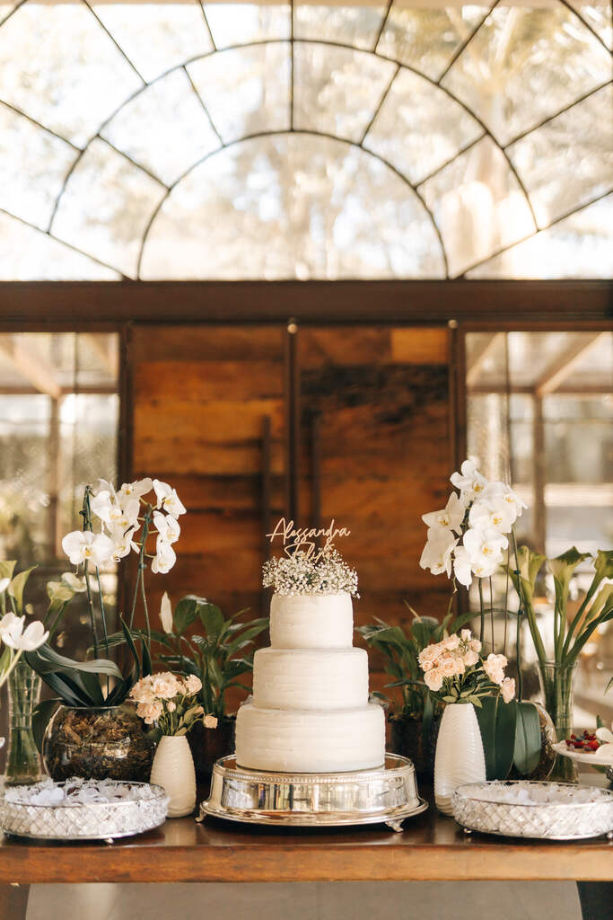 mesa de madeira com bolo de casamento branco com três andares e topo com flores brancas ao lad de orquideas brancas