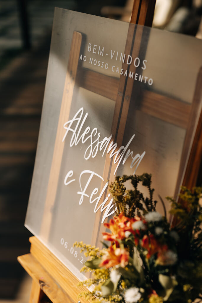 cavalete de madeira com quadro de acrilico semi transparente com o nome dos noivos e mini arranjo de flores brancas e laranjas