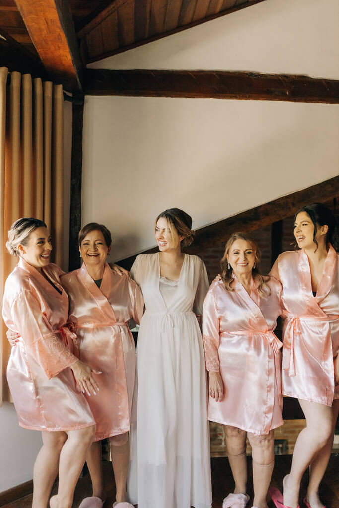 noiva com robe ranco ao lado das madrinhas com com robes rosas claro acetinados dentro de quarto rústico