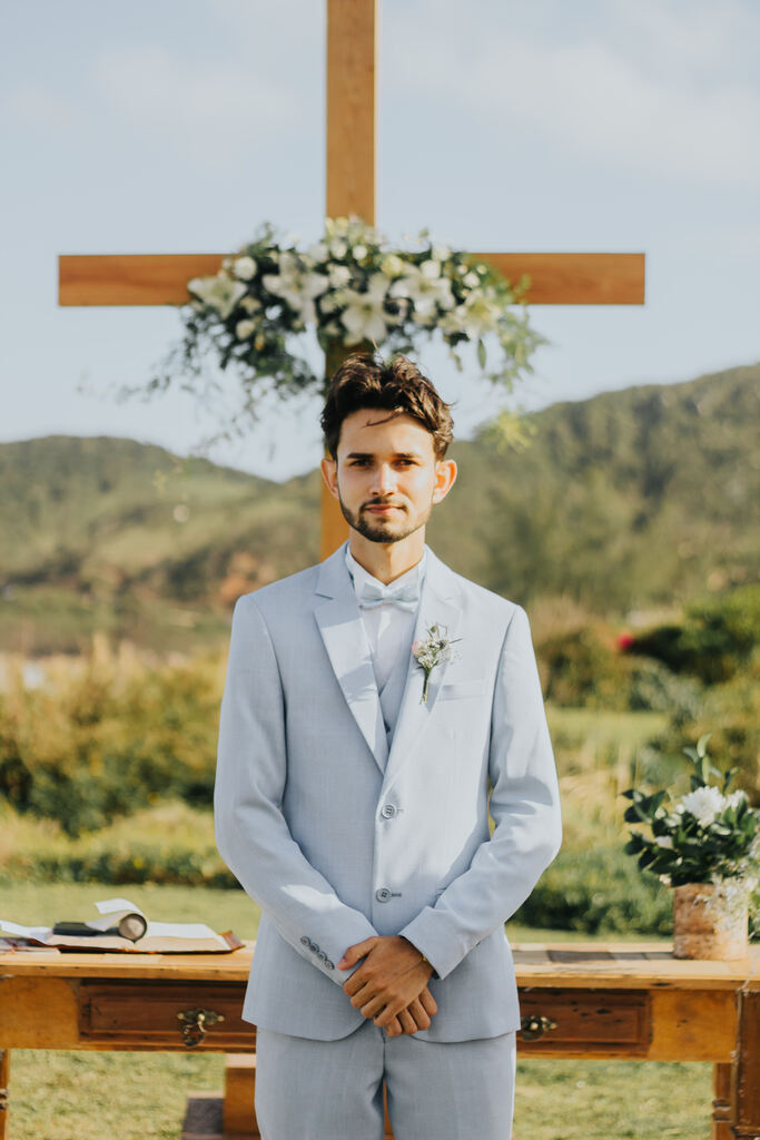 noivo com terno azul claro na frente do altar em forma de cruz de madeira