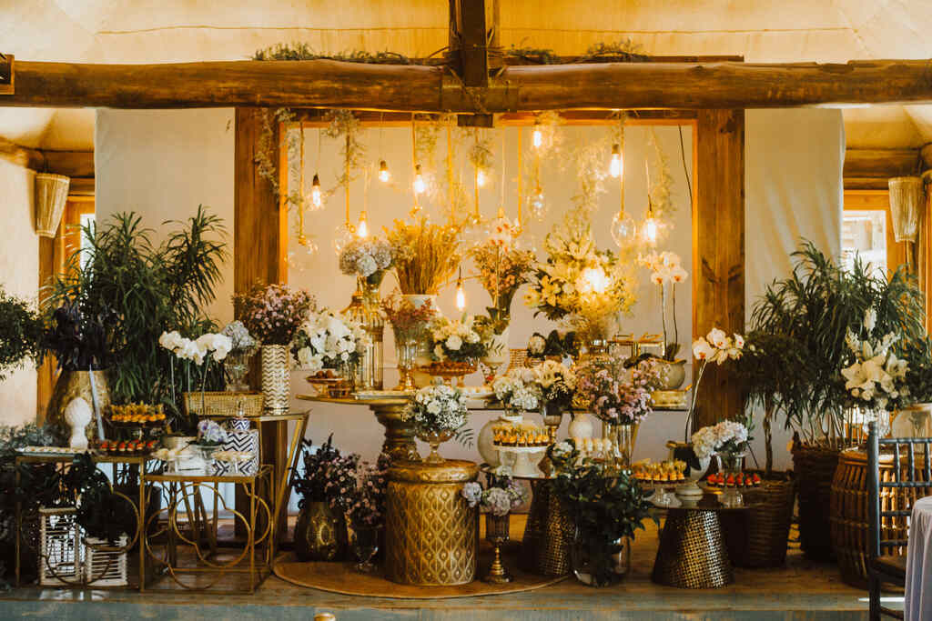 salão com mesas de madeira com arranjos com flores e platas e luminárias pendentes