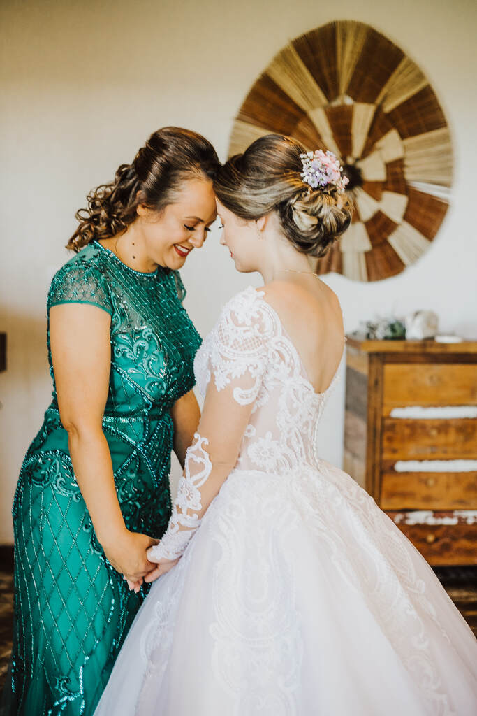 mãe da noiva com vestido verde com bordado e brilhante de mãos dadas com a noiva com vestido com decote em v nas costas e cabelo preso