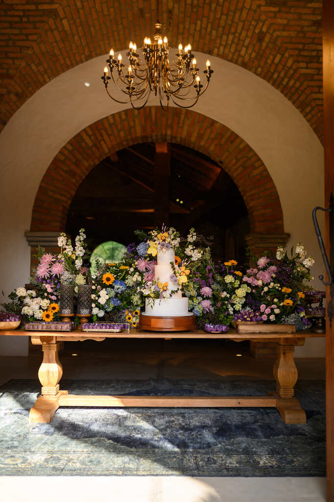 mesa de madeira com flores lilás e girassois em torno do bolo de casamento branco com flores
