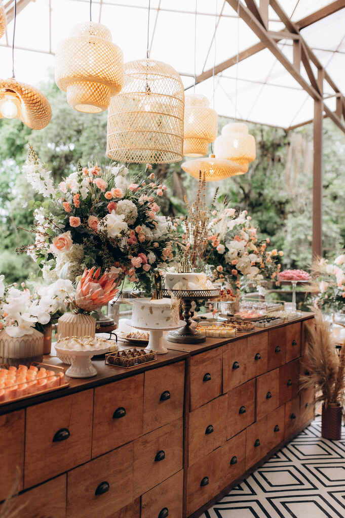 mesa de buffet com bolos de casamento e ao lado de vasos cm flores brancas e rosas e luminárias de palha pendentes