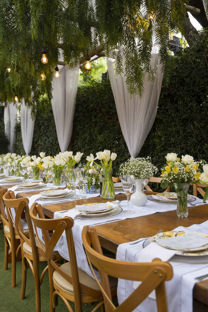 mesas de madeiras posta com tecido branco arranjos com arranjos com flores brancas amarelas e azuis