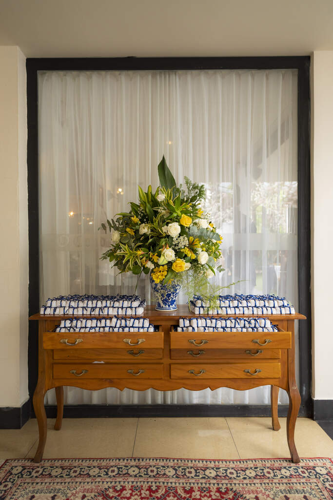 mesa de madeira com bem-casados brancos com laços azuis e vaso no centro com flores brancas e amarelas
