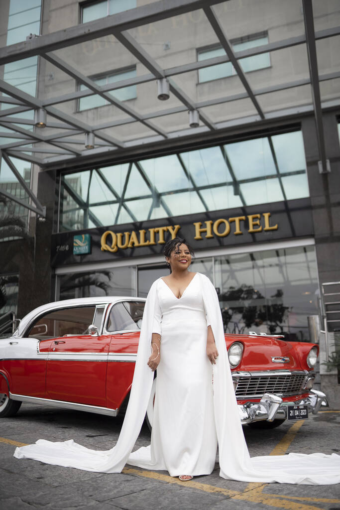 noiva com vestido com decote em v manga longa e capa lateral na frente de carro antigo vermelho e branco na frente de hotel