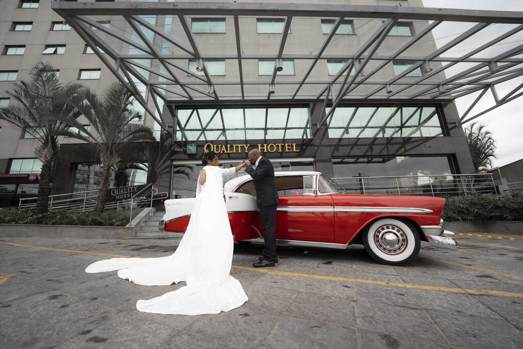 homem com terno preto beijando a mão da noiva com vestido com decote em v manga longa e capa lateral na frente de carro antigo vermelho e branco na frente de hotel