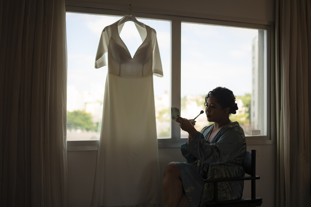 mulher com robe azul sentada na cadeira se maquiando ao lado de vestido de noiva pendurado na frente da janela