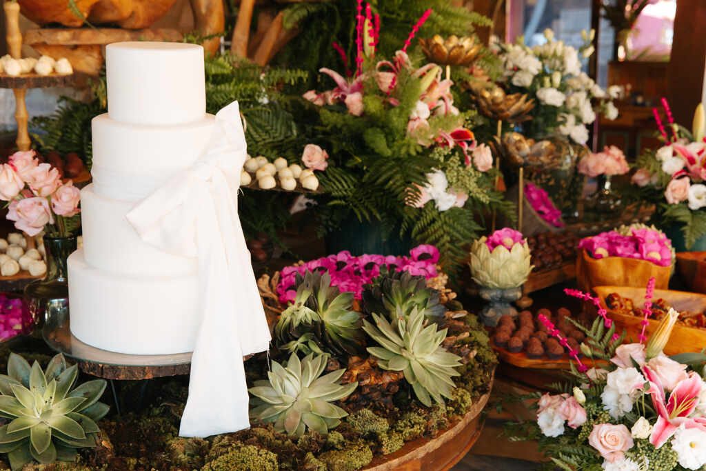 bolo de casamento com quatro andares com laço ao lado de mesa com suculentas e flores brancas e rosas