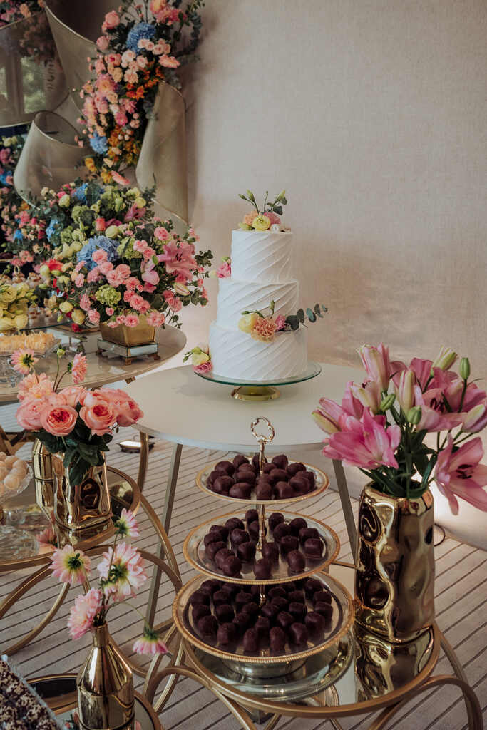 mesa redonda branca com bolo de casamento branco espatulado com topo de flores coloridas e ao lado vários vasos de flores e bandeja com doces de chocolate