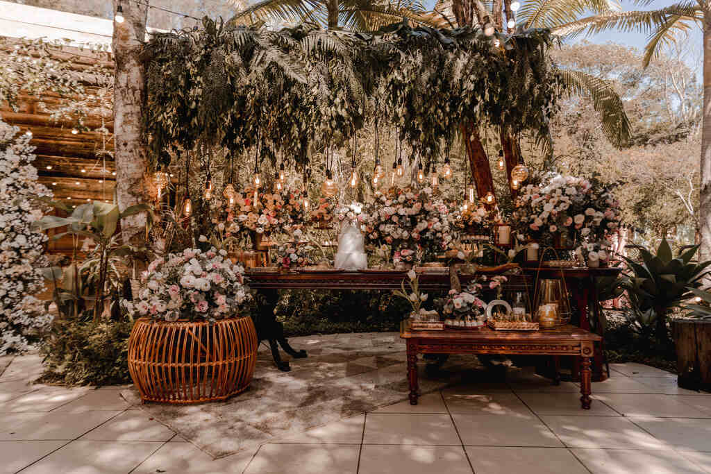 mesa de madeira com bolo de casameto ao lado de vasos com flores brancas e rosas e luminárias pendentes