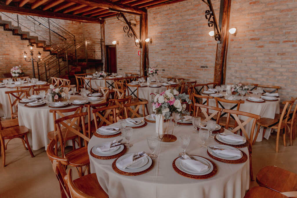 salão rústico ao lado de mesas redondas com toalhas brancas sousplat de palha e flores brancas e rosas no centro