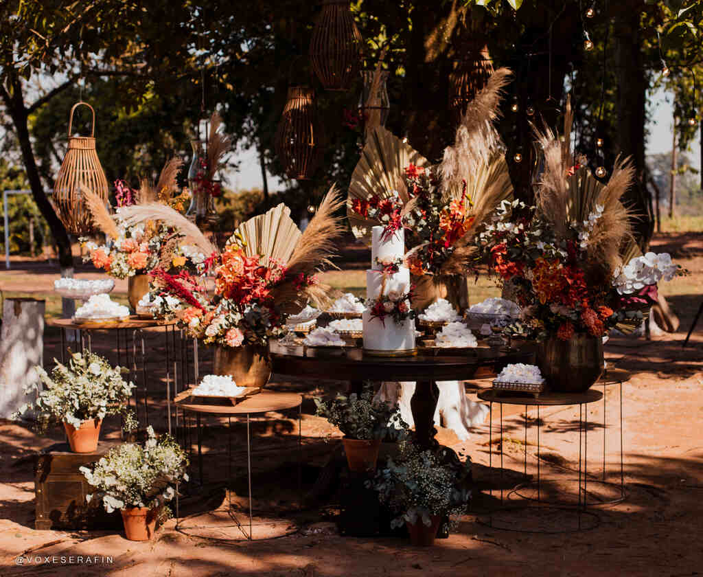 mesa de madeira com bolo de casamento branco com topo com flores e ao lado vasos de barro com flores vermelhas e laranjas e capim dos pampas