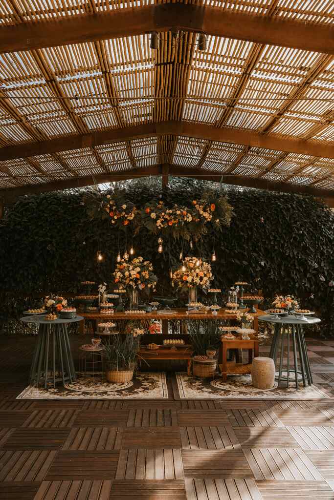 salão com mesa de madeira com três bolos de casamento doces de casamento e arranjos com flores amarelas brancas e laranjas e dois tapetes