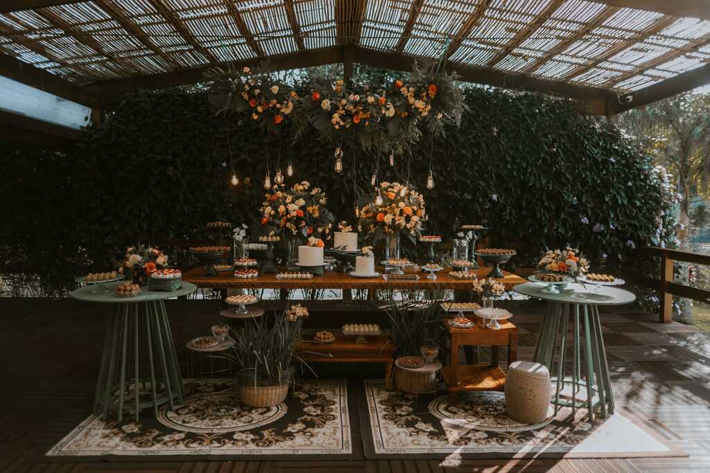 salão com mesa de madeira com três bolos de casamento doces de casamento e arranjos com flores amarelas brancas e laranjas