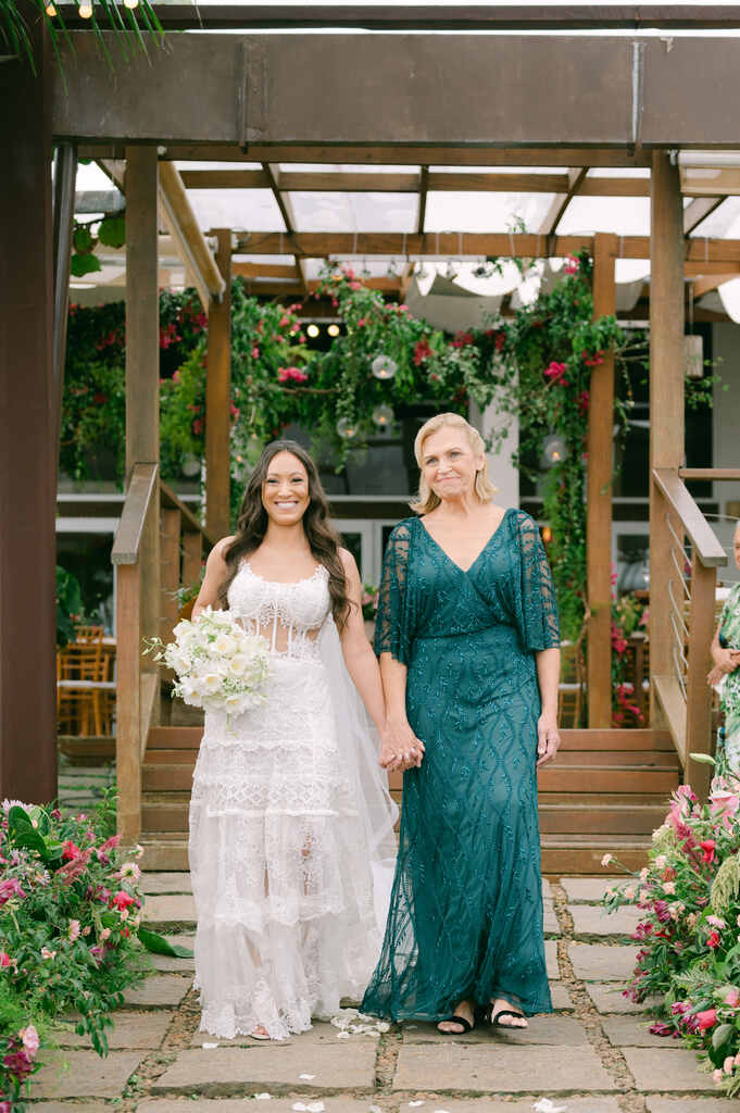 entrada da noiva com vestido com camadas bordadas segurando buquê branco de mãos dadas com amãe com vestido longo verde com decote em v