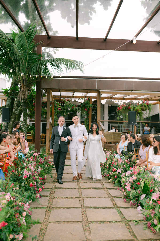 entrada do noivo com terno off white com sapato mrrom ao lado do pai com terno preto e mãe com vestido com capa cinza claro