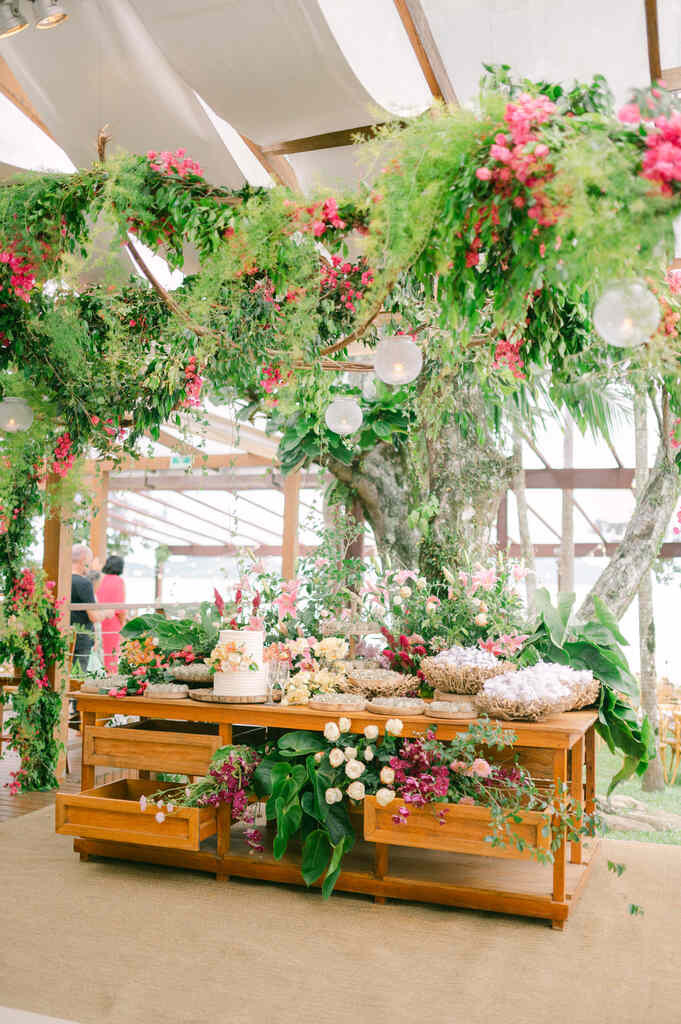 mesa de madeira com bolo de casamento ao lado de flores e plantas e bandejas com doces de casamento