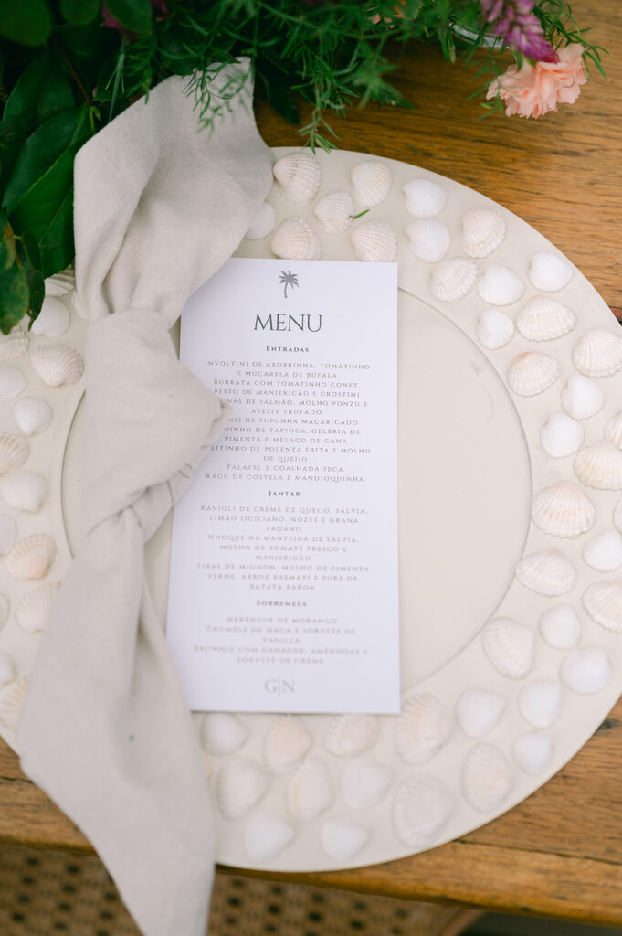 prato com detalhes de conchinhas ao lado com guardanapo com nó e menu para casamento branco