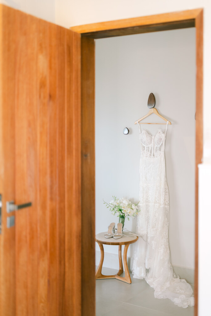 vestido de noiva pendurado no cabide de madeira ao lado de mesinha com flores no quarto branco