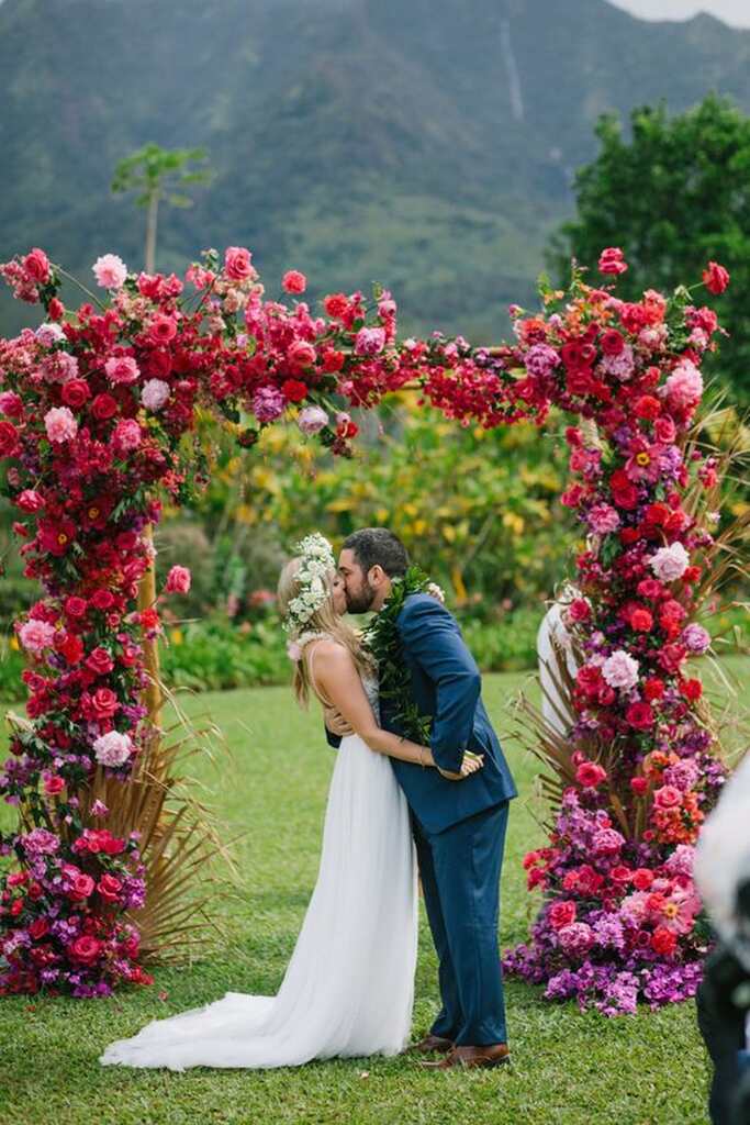 noivos se beijando sob altar quadrado com flores na cor fucsia e vermelho