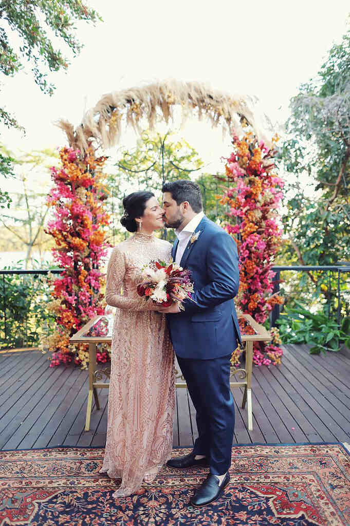 noiva com vestido bordado rosê segurando buquê ao lado do noivo com terno azule ao fundo com altar floral